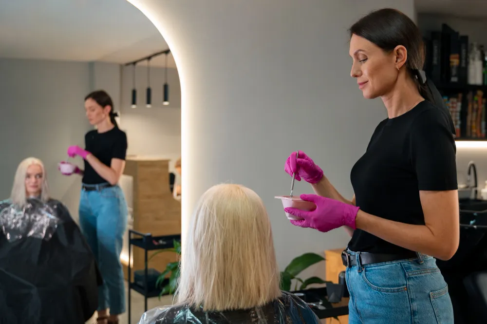 Stilist profesionist aplicând vopsea blondă unei cliente într-un salon modern de înfrumusețare și coafură.