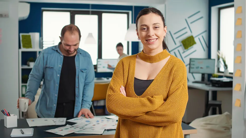 Antreprenoare zâmbind cu brațele încrucișate în biroul unei întreprinderi mici și mijlocii, în timp ce colegii lucrează în fundal