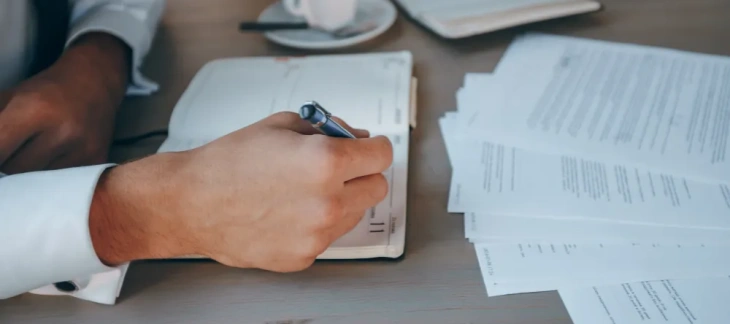 Antreprenor care notează cheltuieli într-o agendă, lângă un teanc de documente și o ceașcă de cafea