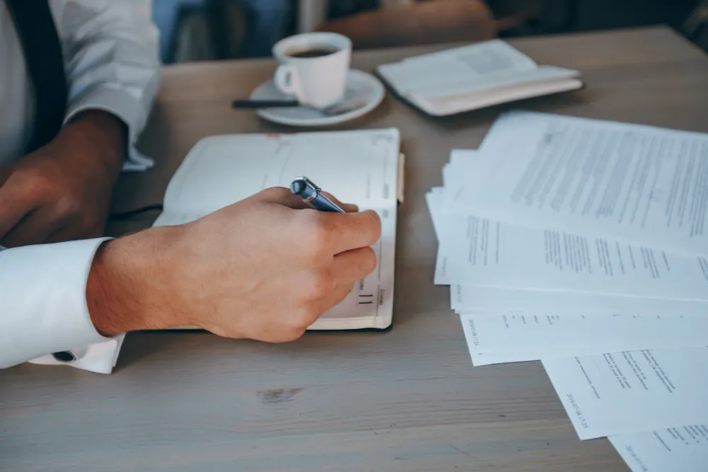 Antreprenor care notează cheltuieli într-o agendă, lângă un teanc de documente și o ceașcă de cafea